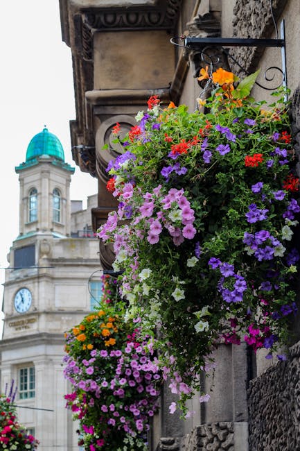 Flower arrangement featuring a vibrant cascade of petunias, geraniums, and marigolds in shades of purple, pink, red, orange, and white, displayed in hanging baskets mounted on a historic stone buildin