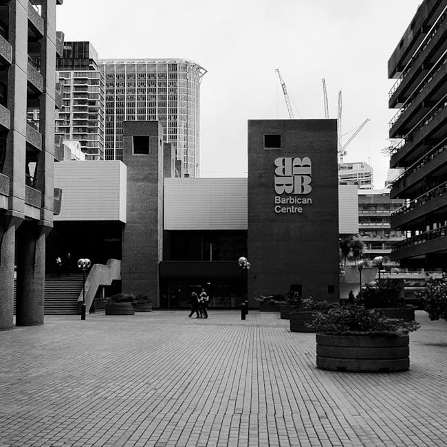 A black and white photograph of the Barbican Centre in London, showcasing its modern architectural design with tall, rectangular brick structures featuring large windows and the prominent 'Barbican Ce