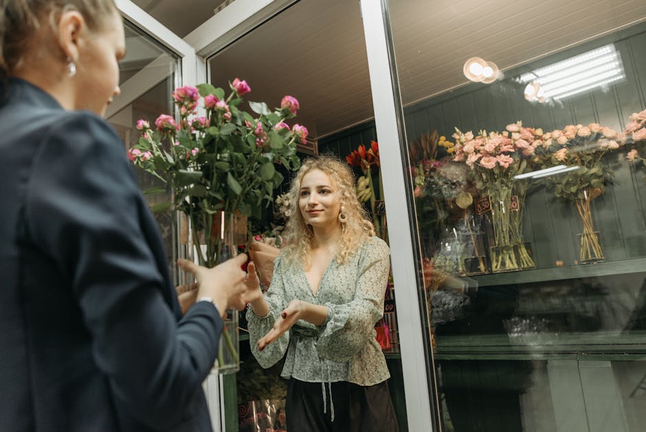 A young woman with curly blonde hair, wearing a light floral blouse, is inside a flower shop, receiving a bouquet of pink roses with green stems and leaves from a florist dressed in a dark jacket. The