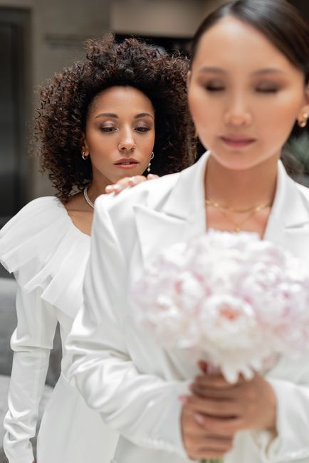 A bride in a white wedding dress with ruffled details on the shoulders holds a bouquet of pink and white flowers, wrapped in light pink paper. She has dark hair styled neatly and wears subtle jewelry.
