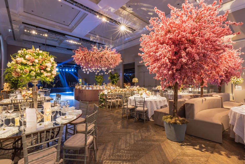An elegantly decorated event space featuring multiple floral arrangements, including a large pink blossoming tree in a grey pot positioned beside a beige curved sofa, and several tall vases filled wit