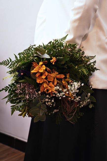 A person dressed in a white blazer and dark skirt is holding a large, lush bouquet of fresh flowers. The arrangement features a variety of vibrant and muted tones, including orange lilies, delicate wh