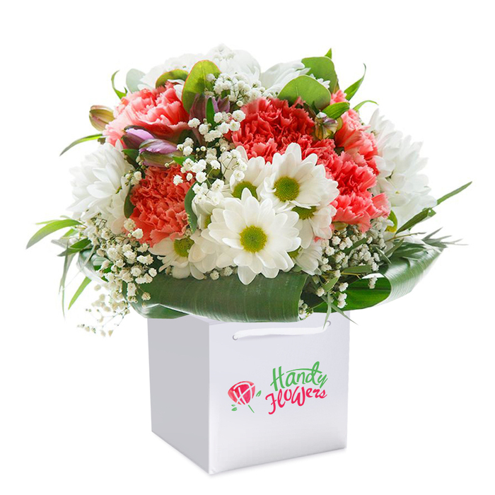 In the luminous light typical of Barbican mornings, a generously sized bouquet rests upright in a clean, contemporary square white gift bag, its pure palette echoing the famous architecture just steps away. Plump ruffled carnations in shades of coral, peach, and vibrant red fill the centre, their velvety petals layered like miniature works of art. Intermixed are daisy-like chrysanthemums with crisp white petals framing chartreuse-lime centers, their cheerful look reminiscent of spring meadows along Golden Lane. Wisps of delicate white gypsophila create a floating, airy feel throughout, while unopened buds with hints of soft purple catch the observer's eye and add expressive depth to the display. Encircling all is a collar of glossy, deep green foliage, providing structure and a rich natural contrast. The bouquet emerges from a stylish white gift bag with sturdy ribbon handles-ideal for gifting or delivering to Barbican residences or offices. The scene's professionally lit simplicity lets the colours and textures shine, highlighting the freshness and care behind each floral choice. Whether destined for a joyful birthday, thoughtful thank-you, or a milestone celebration within EC2, this arrangement makes a refined statement about the enduring beauty of expertly crafted flowers from a trusted local Barbican florist.