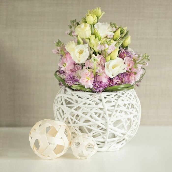 Set against the timeless grace of the Barbican's architectural backdrop, this lush floral arrangement resonates with pastel serenity and artisan flair. The bouquet's heart lies in waves of creamy and pale yellow eustoma, their ruffled petals glowing softly under the ambient city light. Wisps of blush pink matthiola rise among them, lending airy elevation, while below, a tapestry of rich purple filler blooms invokes the wild charm of Barbican's planted communal spaces. The surrounding green foliage, deep and glossy, frames the composition, enhancing each bloom's form and freshness. At the base, a striking white vase-robustly spherical, with a detailed open-lattice design woven from painted rattan-elevates the bouquet, its transparent pattern echoing the iconic concrete grids and artistry found throughout Barbican's lanes. On the left, two crafted spheres of pale woven wood repeat the motif, their handmade charm harmonising with the creative spirit of the local district. The table beneath is spotless and white, bathed in soft light that brings out the arrangement's gentle hues, while a subtly textured beige wall behind-suggestive of fine-textured wallpaper favoured in Barbican's modern flats-serves as an elegant backdrop. This sophisticated floral story, shaped by local Barbican expertise, is as fitting for a contemporary wedding or gallery reception as it is for elevating everyday living. The arrangement's graceful lines and rich artistry evoke the enduring connection between nature and urban refinement in the heart of Barbican.