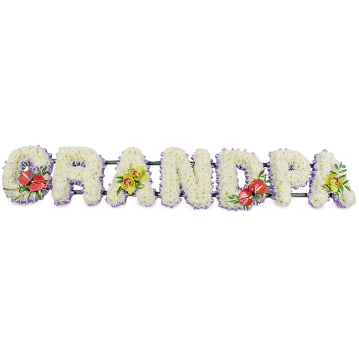 Against a crisp white backdrop, a significant sympathy tribute spells the word "GRANDPA" in broad, block-style lettering, each character brimming with tightly clustered white chrysanthemums. The blooms sit shoulder to shoulder, forming thick, textured cushions that evoke softness and gentle support, much like the emotional comfort family members seek at a time of loss. A slender edging of pale lilac or lavender ribbon wraps the base of every letter, just visible beneath the flowers, tracing a delicate outline around the word and offering a cool, calming hint of colour. Behind the letters, a dark green metal or wooden frame discreetly connects them in a straight line, suggesting that the tribute is ready for display on stands at a funeral or memorial in the Barbican area, perhaps near the arts venues and quiet gardens where families gather to say goodbye. Carefully placed accent sprays bring depth and meaning to the design. The first letter, "G", is embellished with a dramatic cluster of deep red anthuriums, their waxy, heart-shaped bracts and prominent spadices emerging from rich green foliage, conveying passionate remembrance. The initial "A" introduces an elegant spray of yellow-green orchids, their graceful, star-like blooms and glossy leaves adding a note of refined beauty. The "D" echoes the "G" with another grouping of red anthuriums, visually anchoring the centre of the tribute, while the final "A" balances the composition with a matching cluster of orchids. The overall impression is one of dignified tenderness: a custom funeral flower arrangement that would look perfectly at home in a quiet chapel or crematorium near the Barbican, honouring a grandfather's life with tranquillity, respect, and enduring affection.